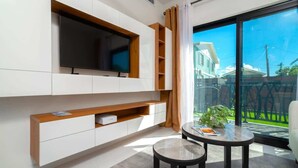Een moderne woonkamer met een flatscreen televisie aan een witte muur, een klein houten tafeltje en een schuifpui naar een balkon.