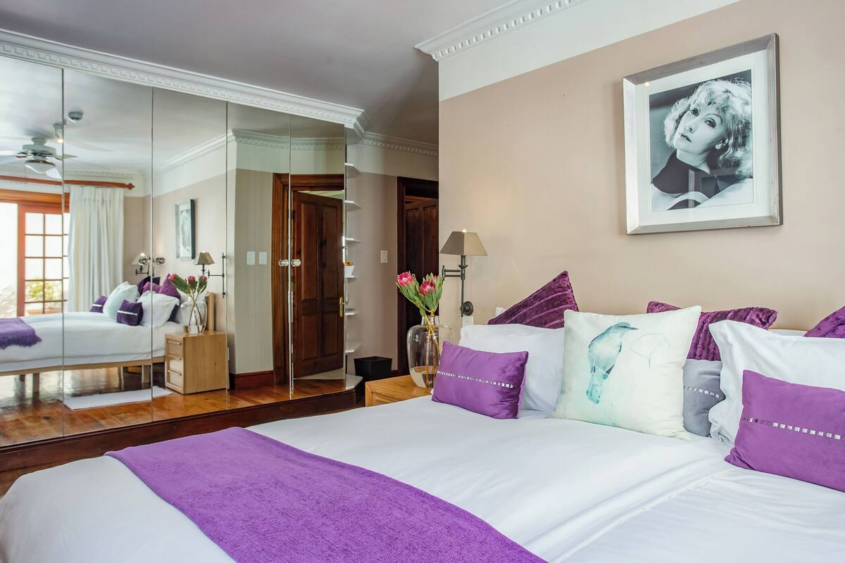 A hotel room with a large bed, a mirror, a framed picture on the wall, and a vase with flowers.
