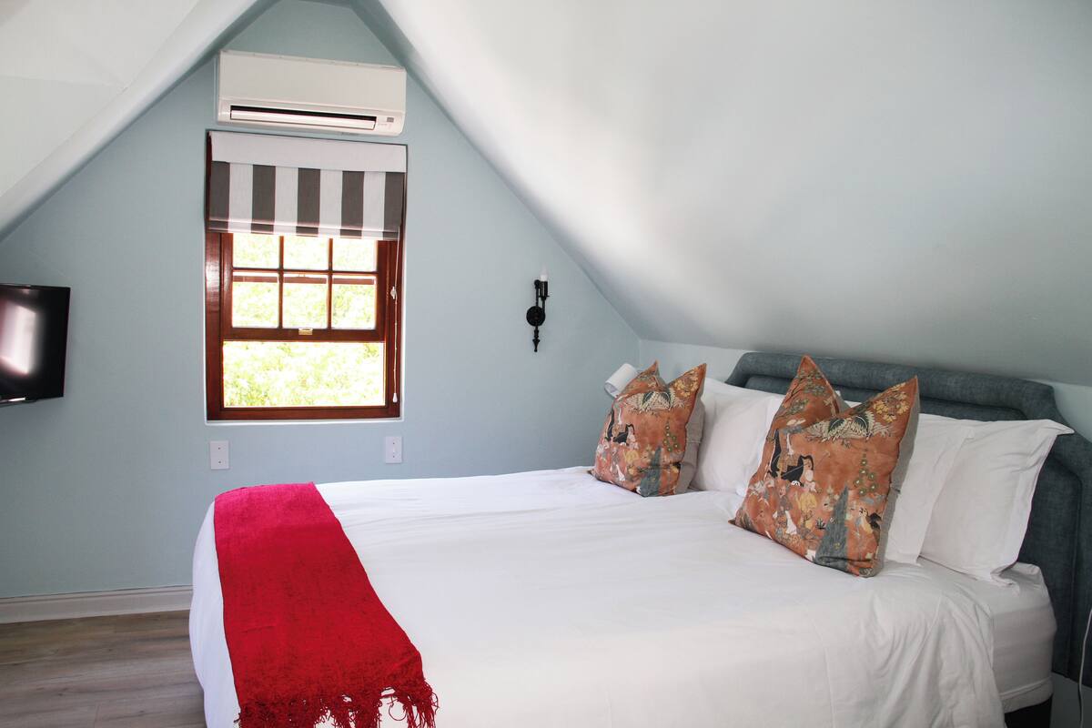 A bedroom with a sloped ceiling, a bed with white bedding and a red throw, two decorative pillows, a window with blinds, and a wall-mounted TV.