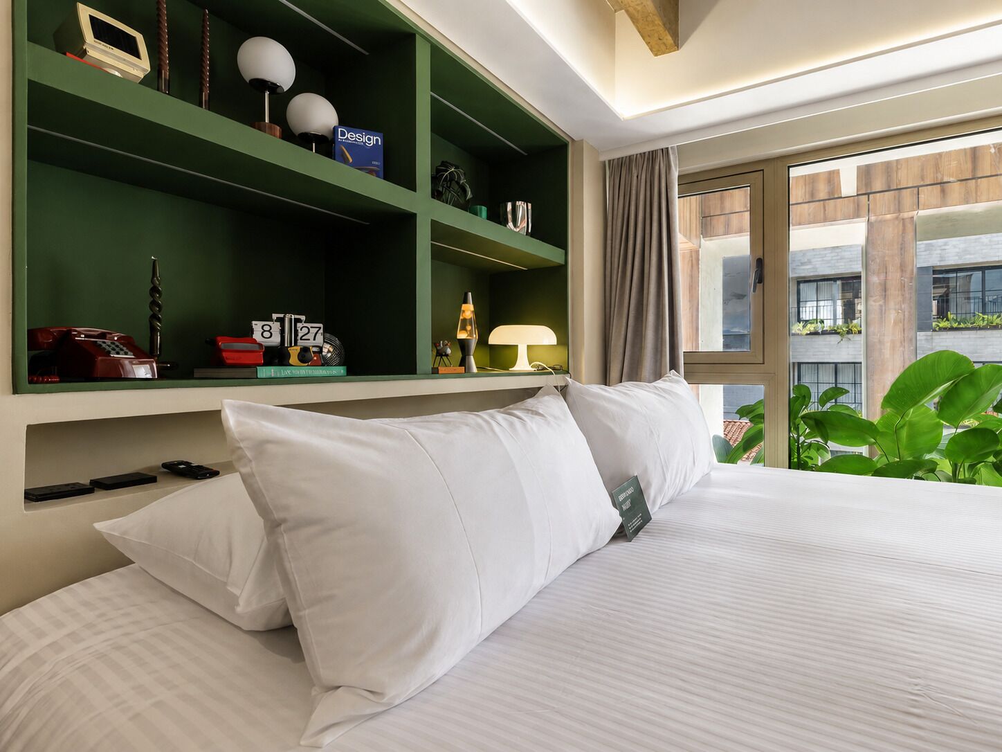 A hotel room with a bed, a green shelf with various items, a window with a view of buildings, and a plant.