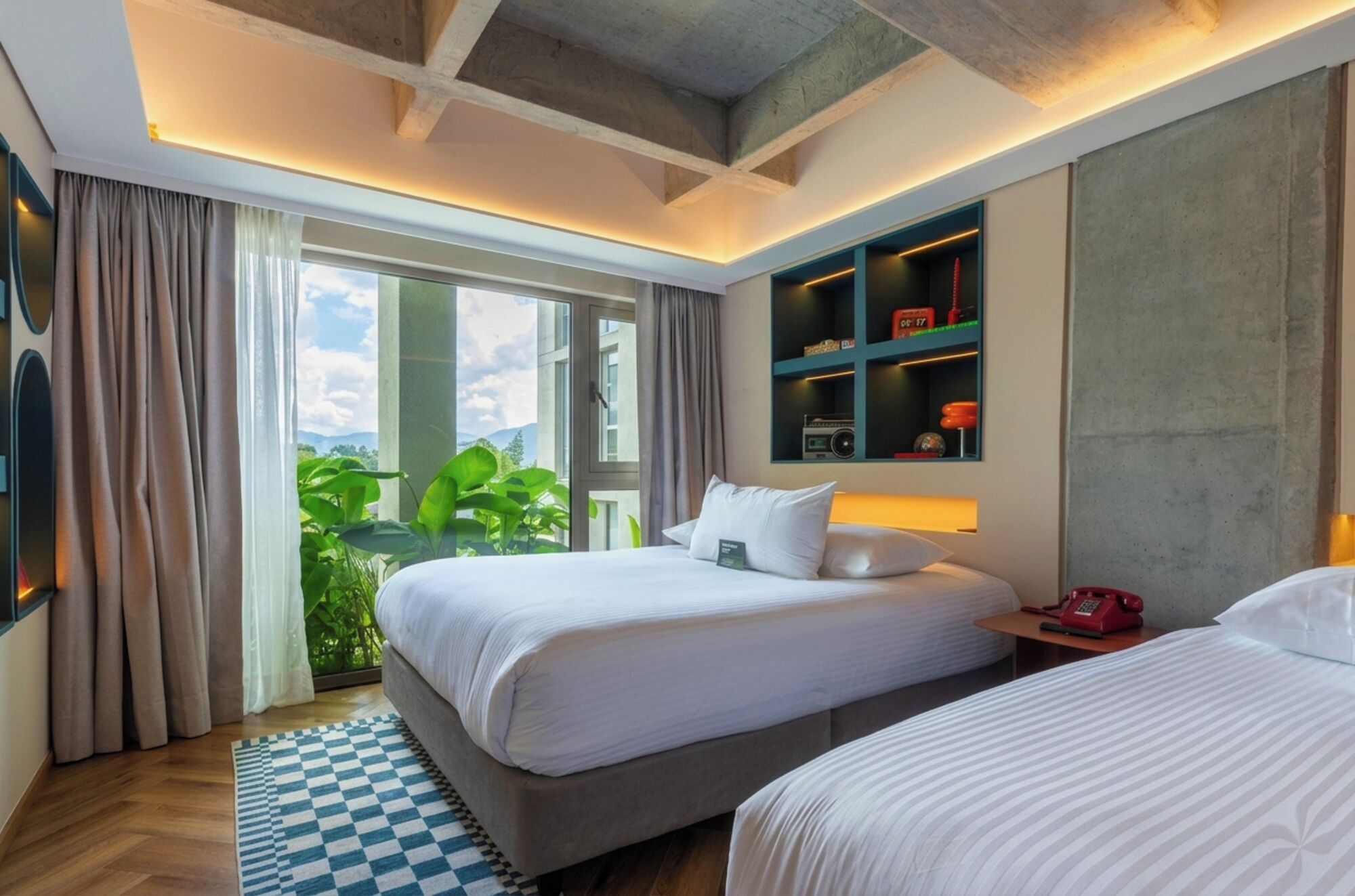 Une chambre d’hôtel avec deux lits, un tapis à carreaux, une grande fenêtre avec des rideaux, une bibliothèque et une vue sur la verdure.
