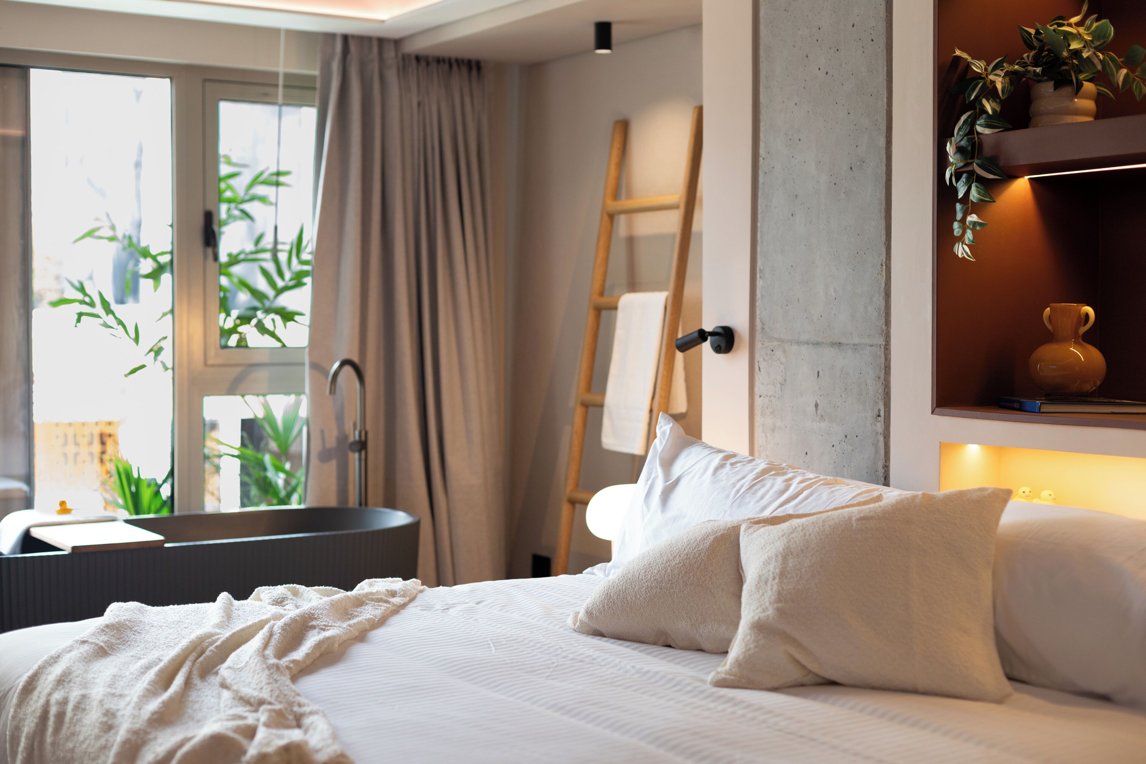 A bedroom with a bed, a window with curtains, a bedside table, a lamp, and a plant.