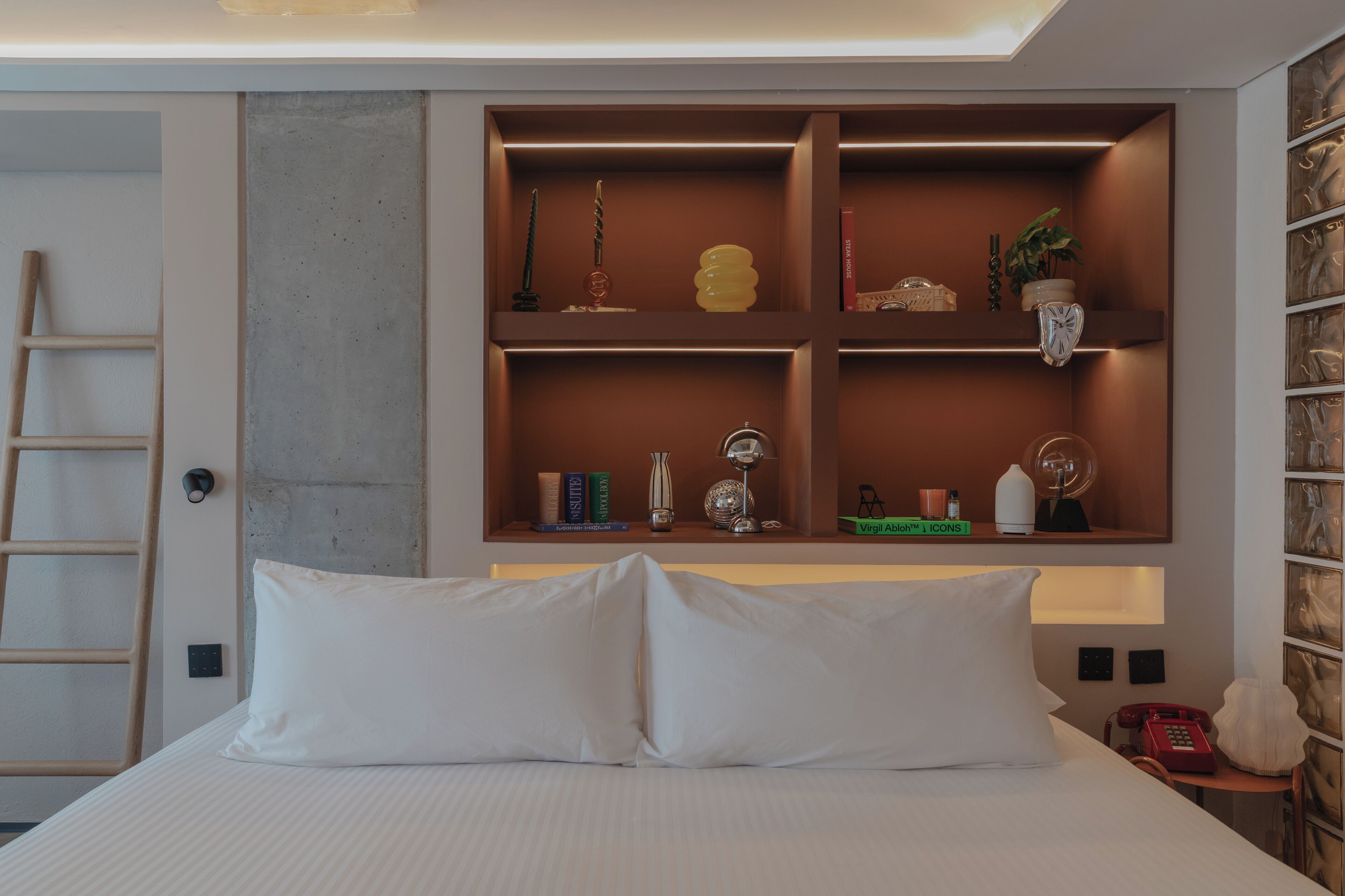 A hotel room with a bed, a shelf with decorative items, a ladder, and a telephone.