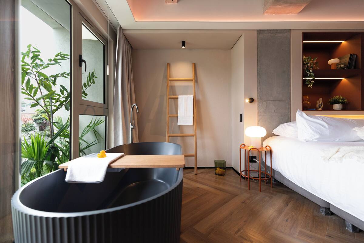 Ein modernes Hotelzimmer mit einer Badewanne, einer Holzleiter, einem Nachttisch mit einer Lampe, einem Bett und Blick auf Grünfläche.