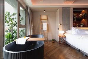 Una camera d'albergo moderna con una vasca, una scala in legno, un comodino con una lampada, un letto e vista sul verde.