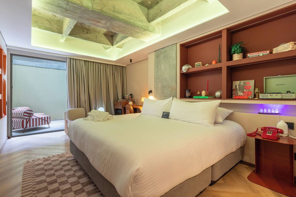 Ein Hotelzimmer mit einem großen Bett, einem Schreibtisch, einem Sessel, einem Regal mit Dekoration und Blick ins Freie.