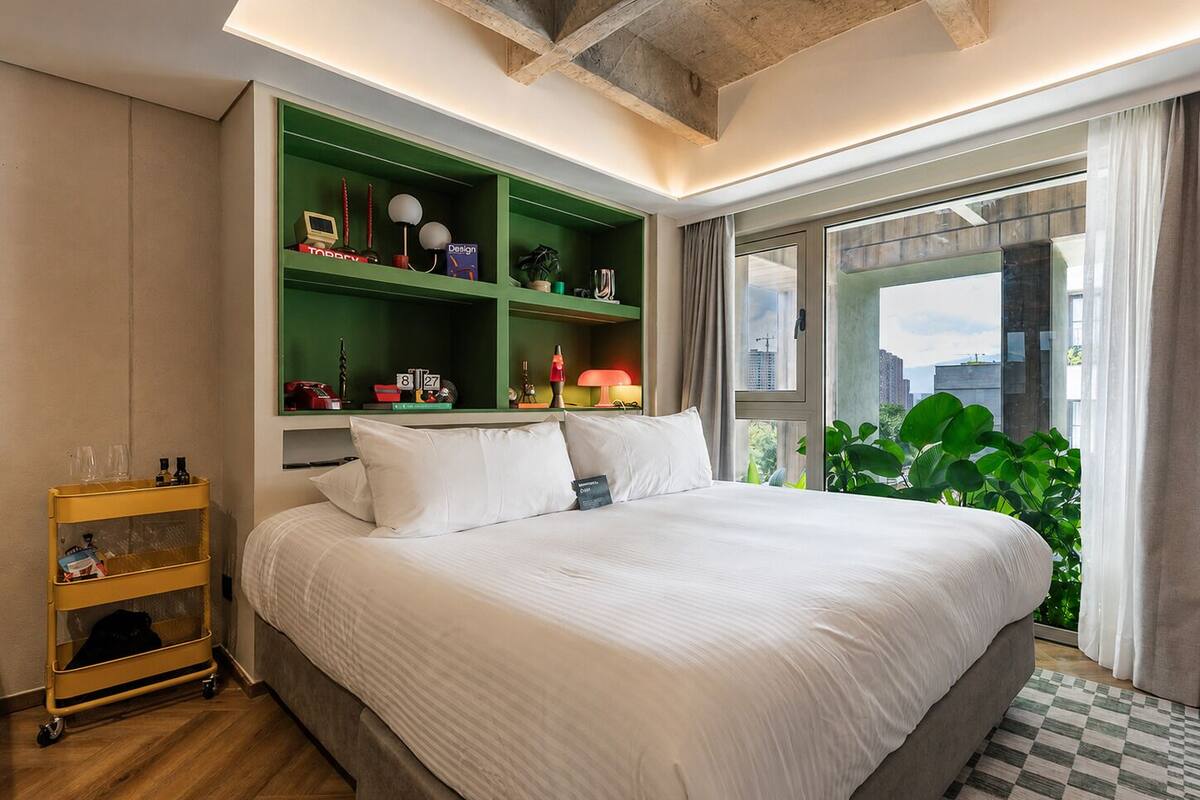 Ein modernes Hotelzimmer mit einem großen Bett, Regalen in Grün mit verschiedenen Gegenständen, einem kleinen Wagen und Blick auf die Stadt durch große Fenster.