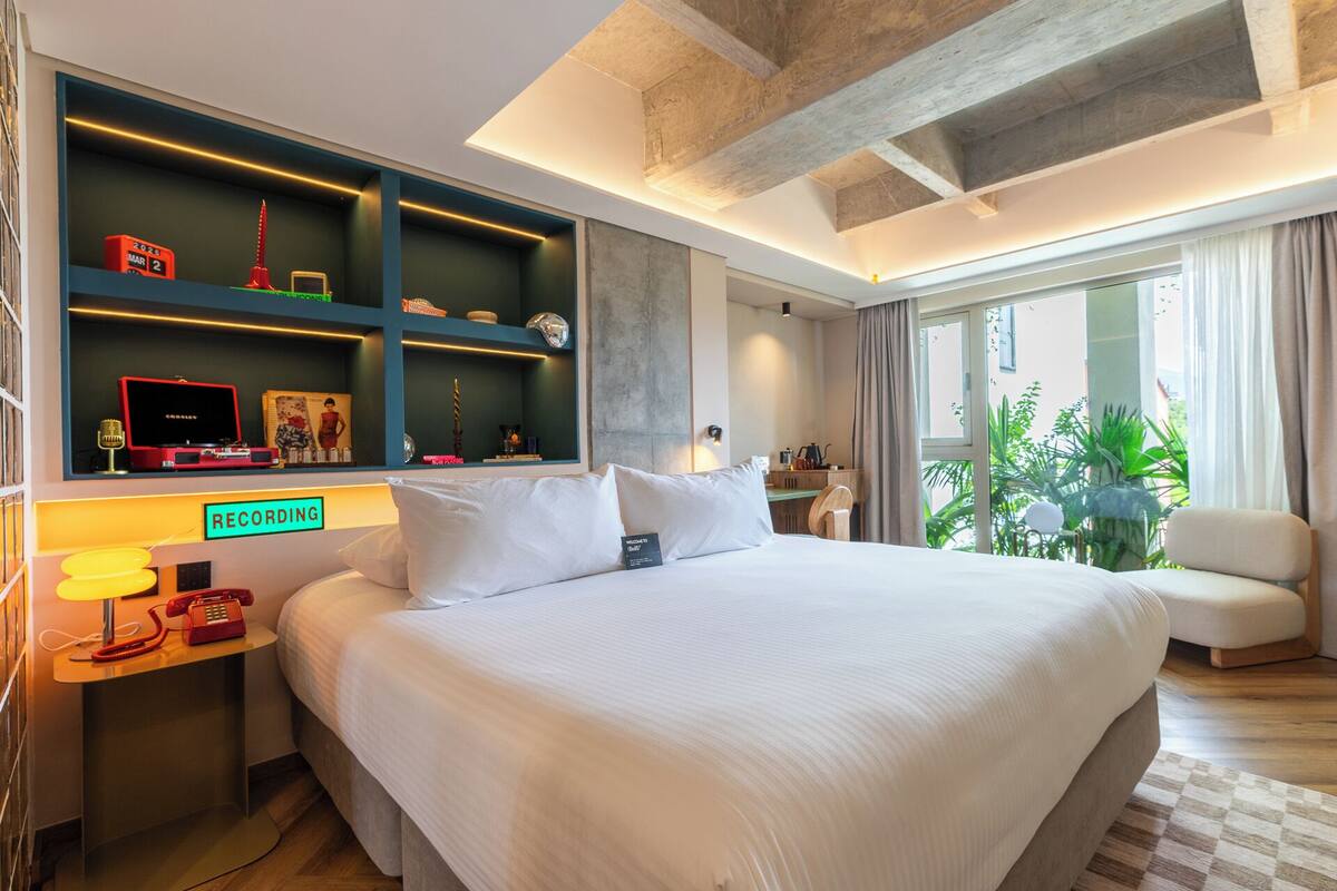 Ein modernes Hotelzimmer mit einem großen Bett, einem Schreibtisch mit Lampe, einem Regal mit Dekoration und Blick ins Freie.
