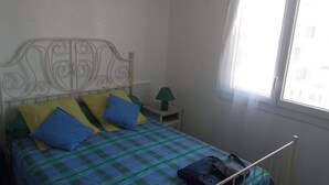 A bedroom with a bed, pillows, a nightstand, a lamp, and a window. - T2 apartment in residence 100 m from the beach. (Canet-en-Roussillon)