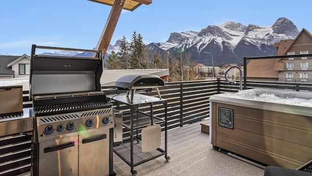 An outdoor patio with a grill, a hot tub, and a view of snow-capped mountains. - Sisters' Solstice - Stunning Mountain Views with Private Hot tub, Sauna & BBQ! (Canmore)