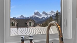 A bathroom with a window view of snow-capped mountains, a modern faucet, and toiletries on the counter. - Sisters' Solstice - Stunning Mountain Views with Private Hot tub, Sauna & BBQ! (Canmore)