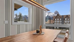 A modern dining area with a wooden table, a candle holder, and a view of snow-capped mountains through large windows. - Sisters' Solstice - Stunning Mountain Views with Private Hot tub, Sauna & BBQ! (Canmore)