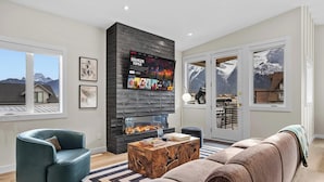 A modern living room with a fireplace, a sofa, a coffee table, and a television mounted on the wall. - Sisters' Solstice - Stunning Mountain Views with Private Hot tub, Sauna & BBQ! (Canmore)