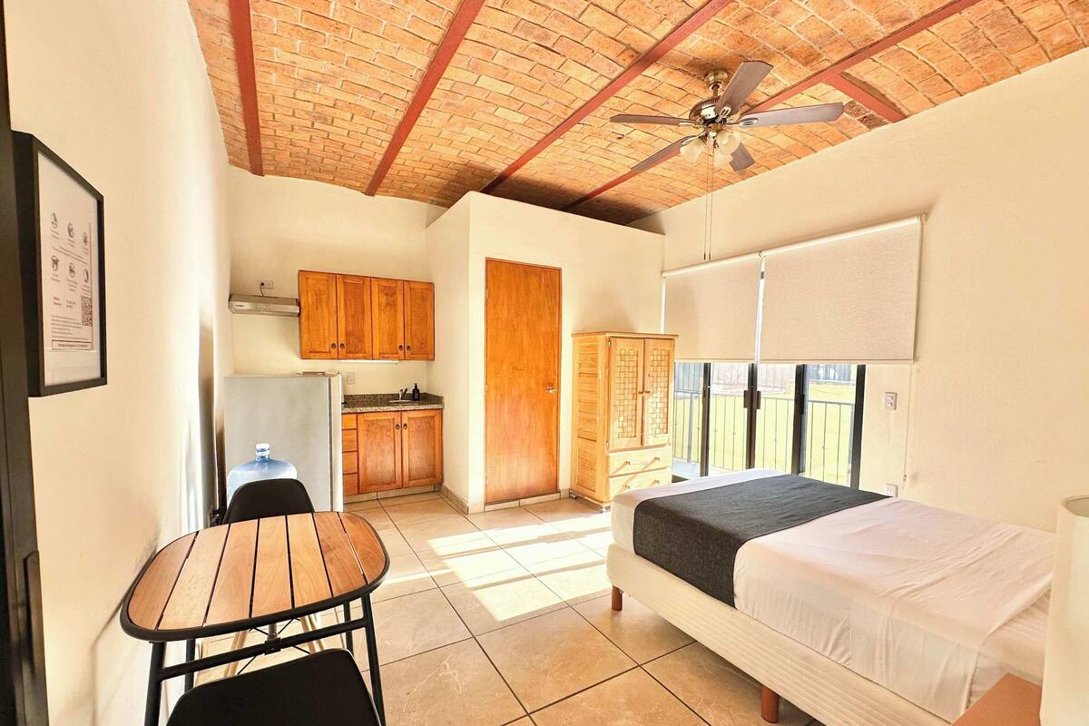 Une pièce de taille modeste comprenant un lit, une petite table, une chaise, un coin cuisine et un ventilateur de plafond.