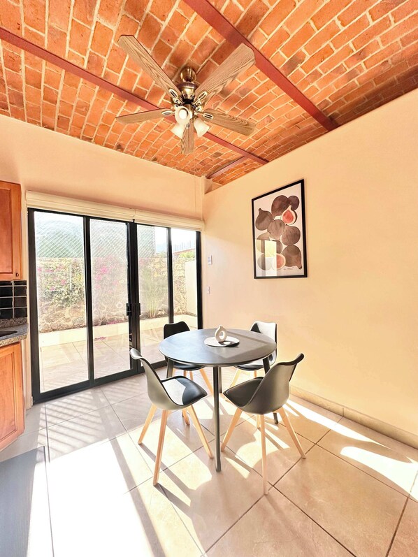 Un coin repas comprenant une table ronde, quatre chaises, un ventilateur de plafond et une grande fenêtre donnant sur un paysage verdoyant.
