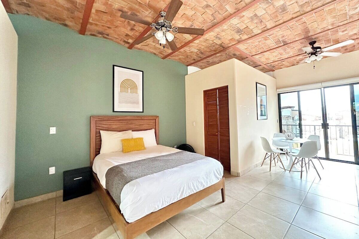 Une chambre à coucher comprenant un lit, un ventilateur de plafond, une tête de lit en bois, un tableau encadré, une fenêtre et un balcon avec une table et des chaises.