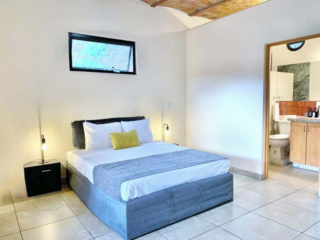 Ein Schlafzimmer mit einem Bett, einem Nachttisch, einem an der Wand montierten Fernseher und einem durch eine offene Tür einsehbaren Badezimmer.
