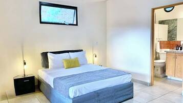 Ein Schlafzimmer mit einem Bett, einem Nachttisch, einem an der Wand montierten Fernseher und einem durch eine offene Tür einsehbaren Badezimmer.