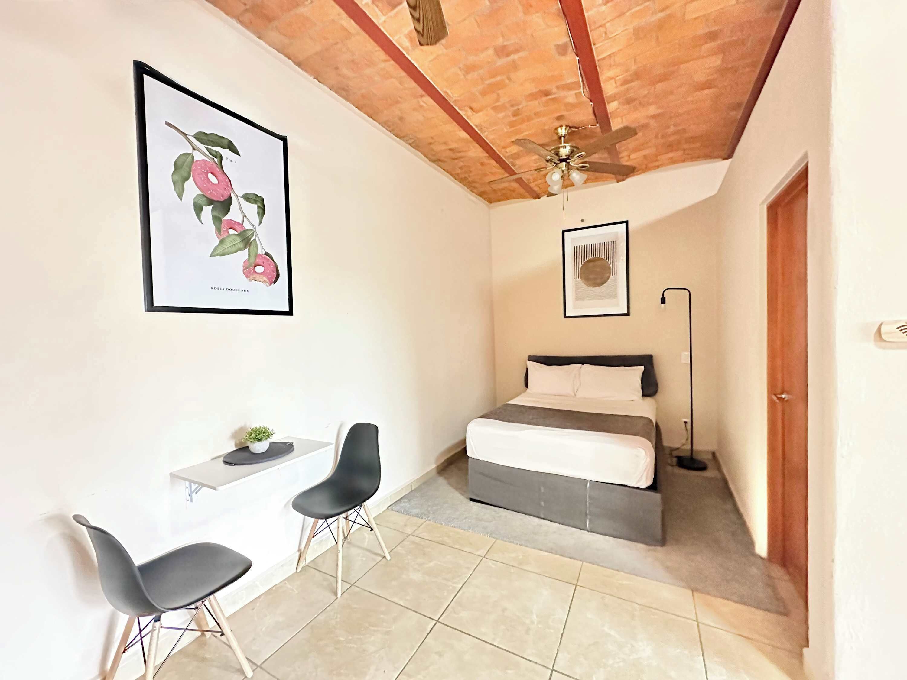 A bedroom with a bed, a desk, two chairs, a ceiling fan, and a framed picture on the wall.