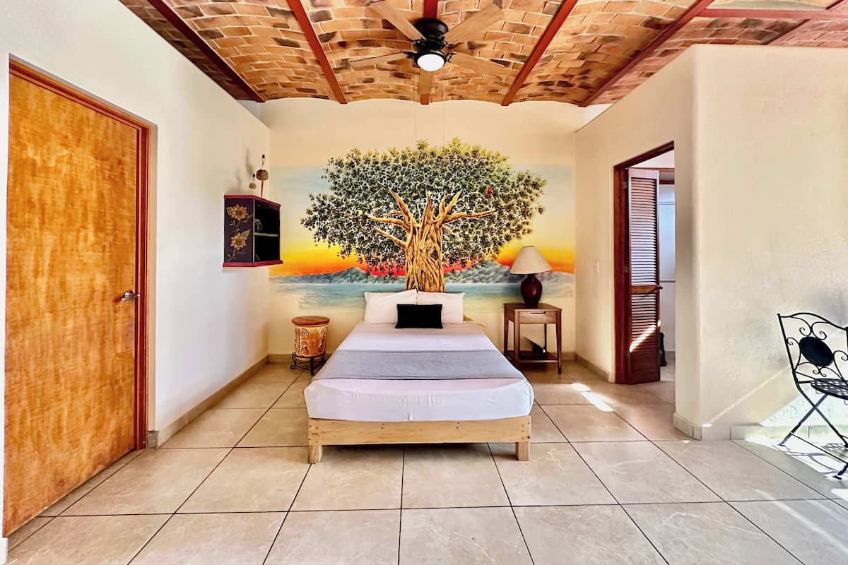 Une chambre avec un lit en bois, un ventilateur de plafond, une fresque représentant un arbre et une vue sur la mer.
