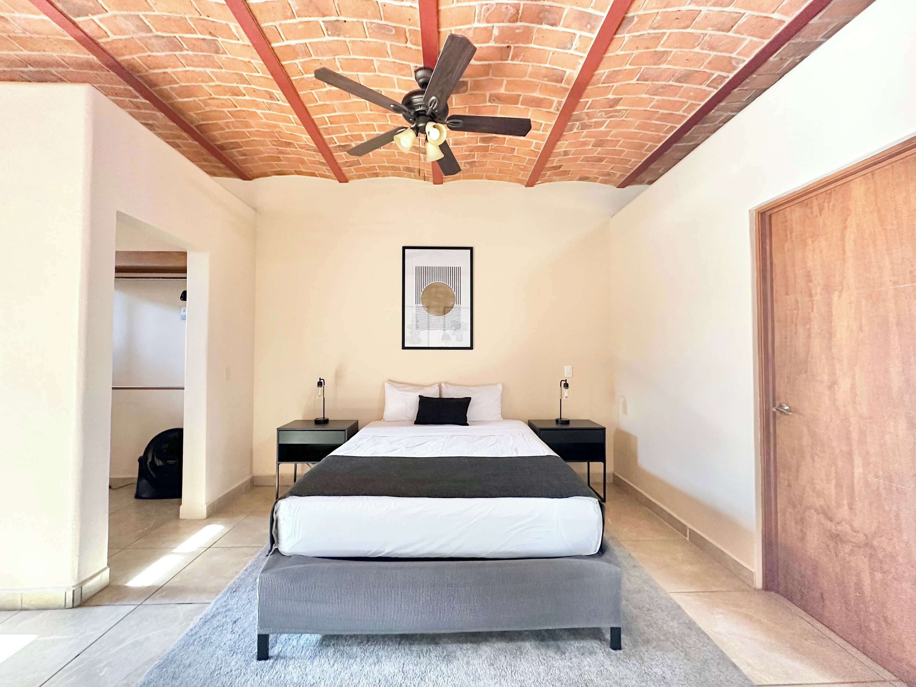 A bedroom with a bed, two bedside tables, a ceiling fan, a framed picture, and a wooden door.