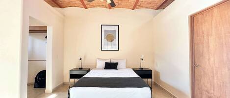 A bedroom with a bed, two bedside tables, a ceiling fan, a framed picture, and a wooden door.