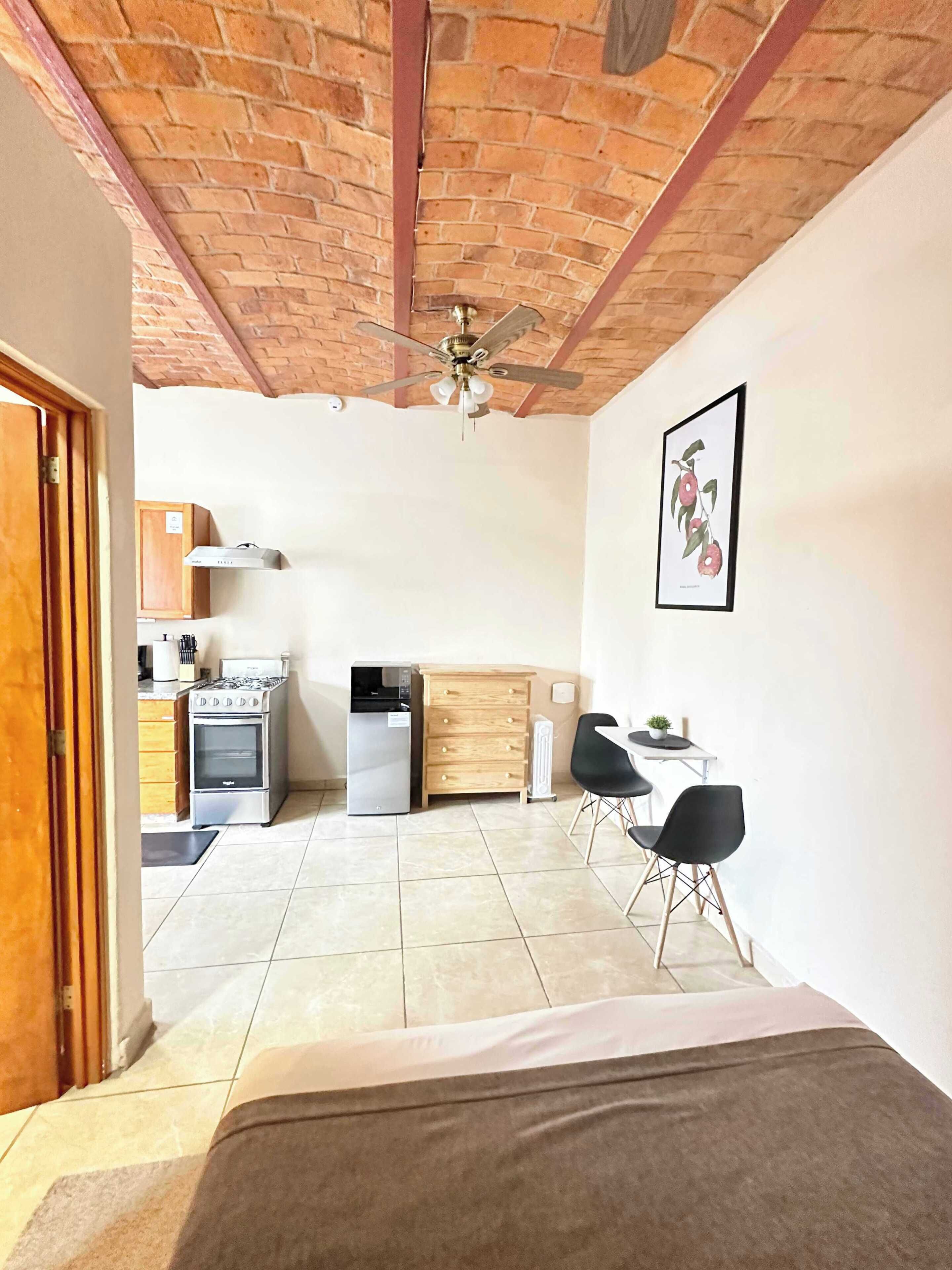 Una habitación funcional con cama, kitchenette, comedor con sillas y ventilador de techo.