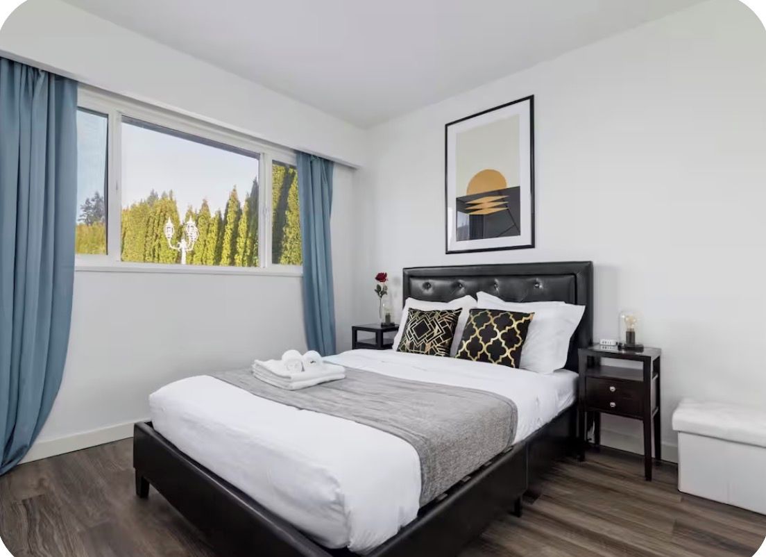 A hotel room with a bed, a nightstand, a bedside table, a window with curtains, and a framed picture on the wall.
