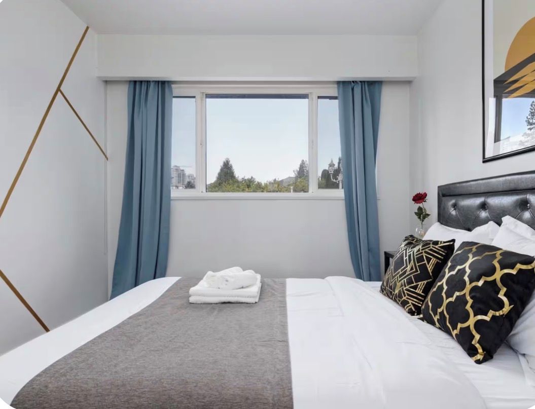 Ein Hotelzimmer mit Bett, einem Fenster mit Vorhängen, Blick auf die Stadt und einem gerahmten Bild an der Wand.