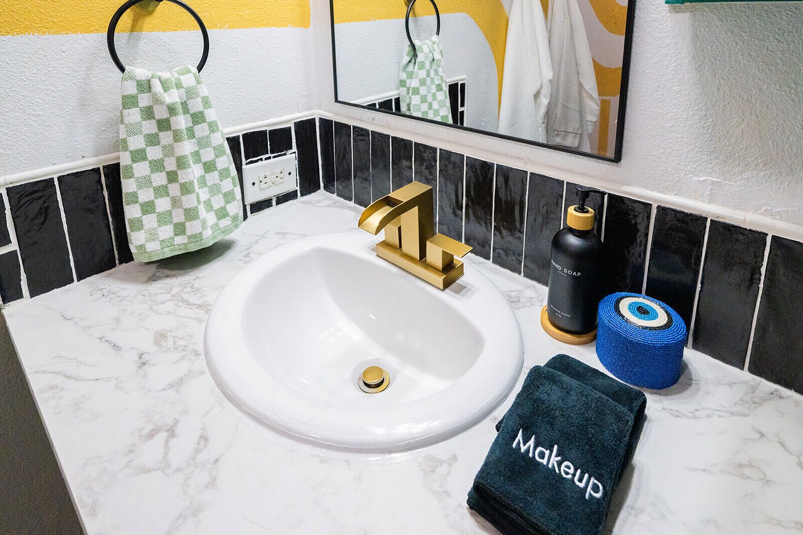Casa de banho com lavatório branco, torneira dourada, salpico de azulejos preto e branco, espelho e porta-toalhas com toalha de padrão quadriculado verde e branco.