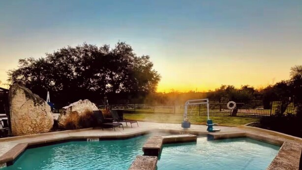 Piscina con borde de piedra y zona de hidromasaje, rodeada de árboles y una valla.