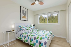 A bedroom with a bed, a bedside table, a ceiling fan, a window with a view of trees, and a framed picture on the wall. - The Forest Pedal House (Squamish)