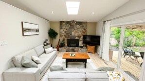 Ein Wohnzimmer mit einer Couch, einem Couchtisch, einem Fernseher, einem Kamin aus Stein und Blick durch große Schiebetüren.