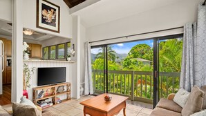 Amplia sala de estar con un televisor de pantalla plana grande, una mesa de centro de madera y un conjunto de sofá. Se ve un balcón con vistas a la vegetación exuberante y montañas a través de las puertas de vidrio correderas.