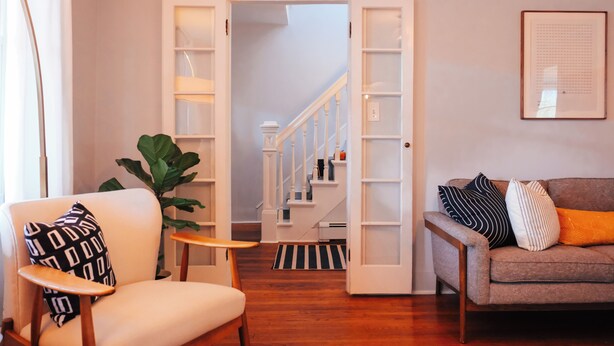 Una sala de estar con un sillón blanco, un sofá gris, una planta verde grande y una mesa de centro de madera. Una escalera con barandillas blancas conduce a un piso superior.