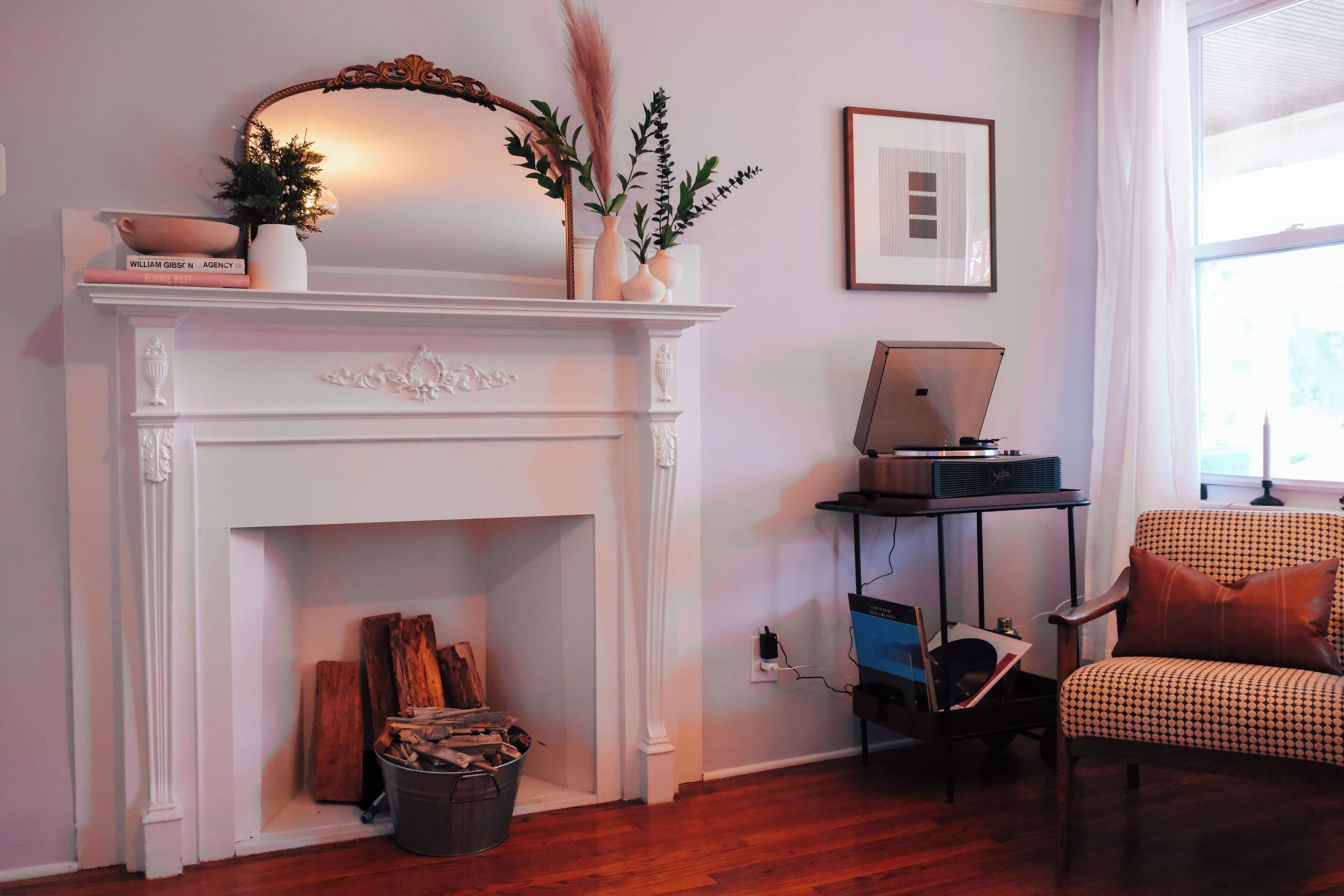 Una sala de estar con una chimenea blanca, una computadora portátil sobre una mesita auxiliar, una silla y un cuadro enmarcado en la pared.
