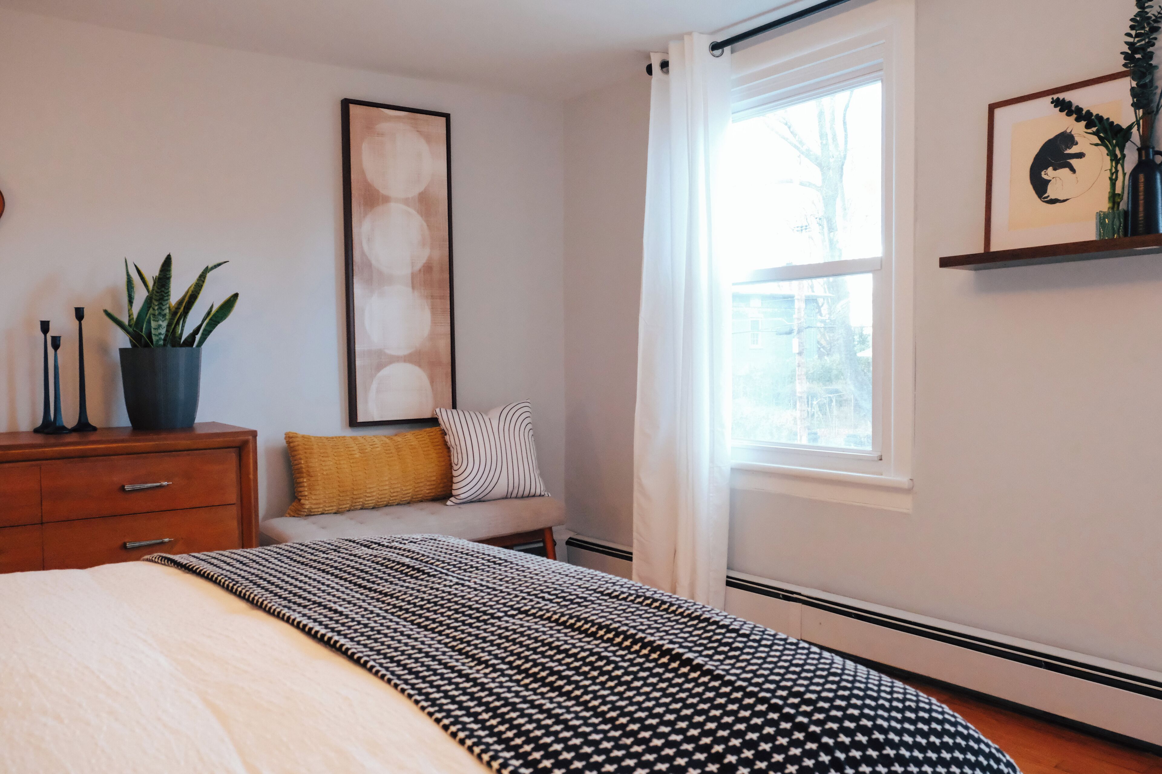 A bedroom with a bed, a bedside table with a plant, a framed picture on the wall, and a window with curtains.