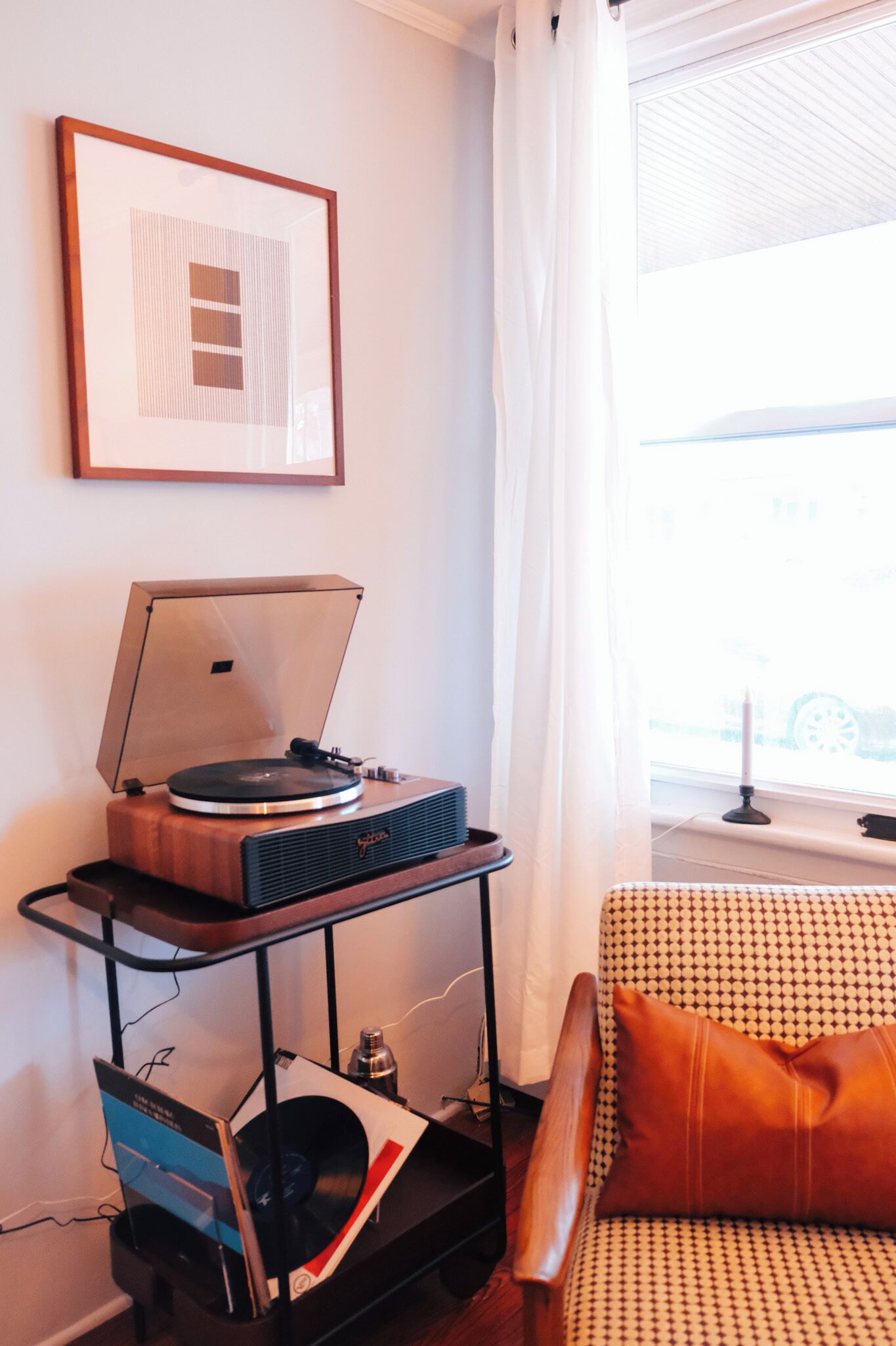 A room with a turntable on a wooden stand, a framed picture on the wall, a patterned sofa, and a window with curtains.