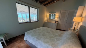 Quarto com uma cama grande, uma janela, mesa de cabeceira com abajur, guarda-roupa e um pequeno banquinho.