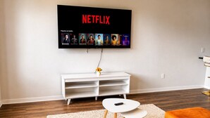 A living room with a flat-screen TV mounted on the wall, a white entertainment unit, a round coffee table, and orange upholstered seating. - Family-Friendly 2BR with Amenities, WiFi & Parking (Des Moines)