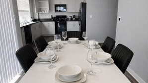 Une table à manger dressée pour six personnes, avec des chaises, une cuisine en arrière-plan et une fenêtre avec des stores.