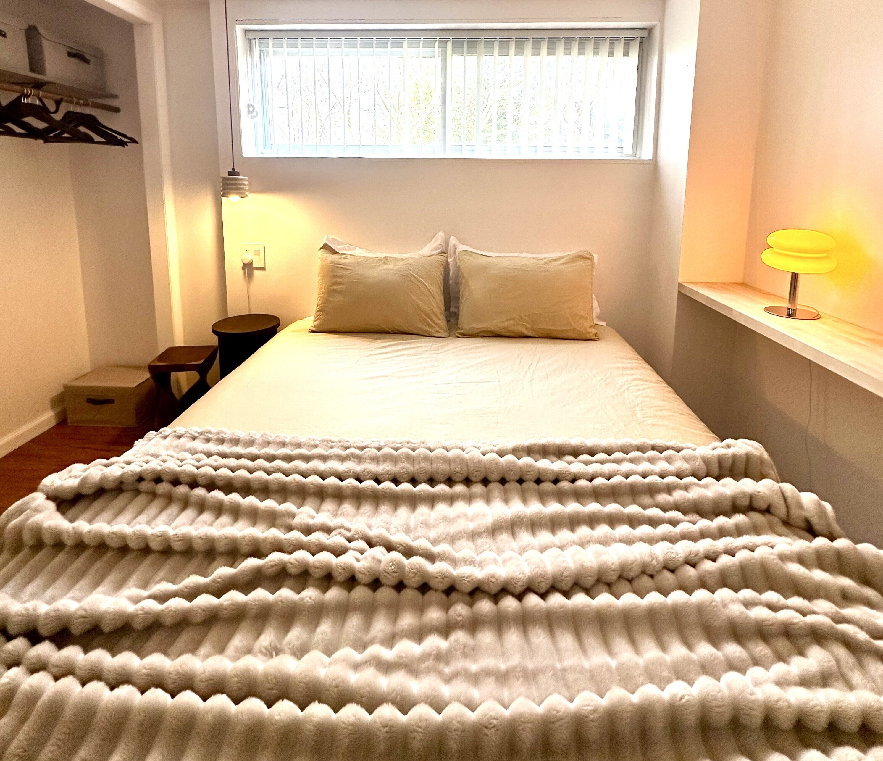 Une chambre à coucher avec un lit, une fenêtre avec des stores, une table de chevet, une lampe et une armoire.