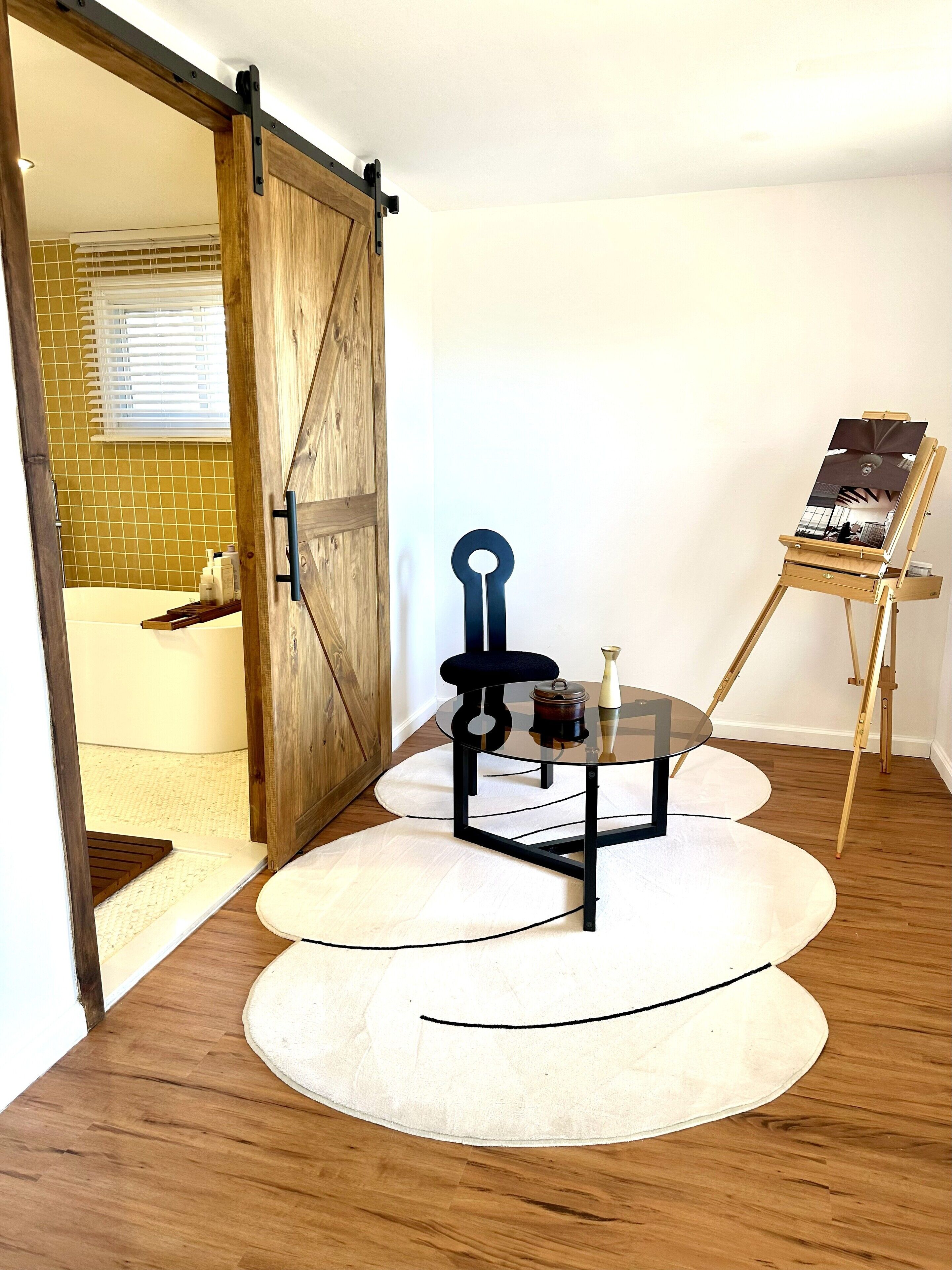 A room with a wooden door, a glass table with a chair, a small table with a vase, and an easel with a painting.