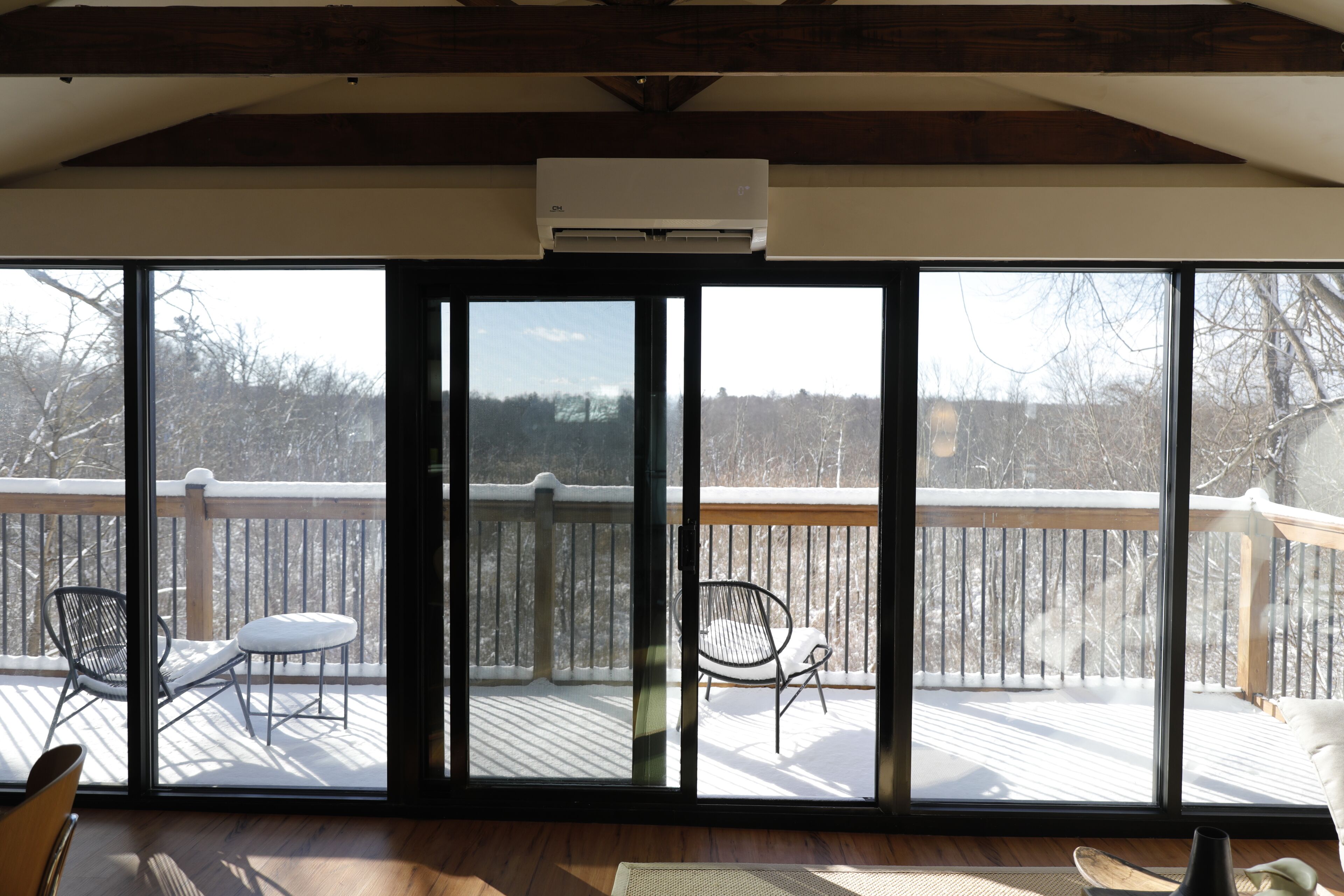 A room with a large sliding glass door leading to a deck with a wooden railing. Inside, there is a wooden floor and a ceiling with exposed beams.