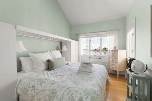Ein Schlafzimmer mit Bett, Kommode, einem Fenster mit Vorhängen und einem Ventilator.