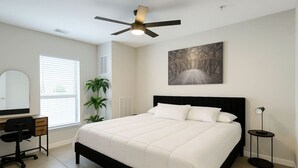 Een slaapkamer met een bed, een bureau, een stoel, een plafondventilator, een vloerlamp en een schilderij aan de muur.