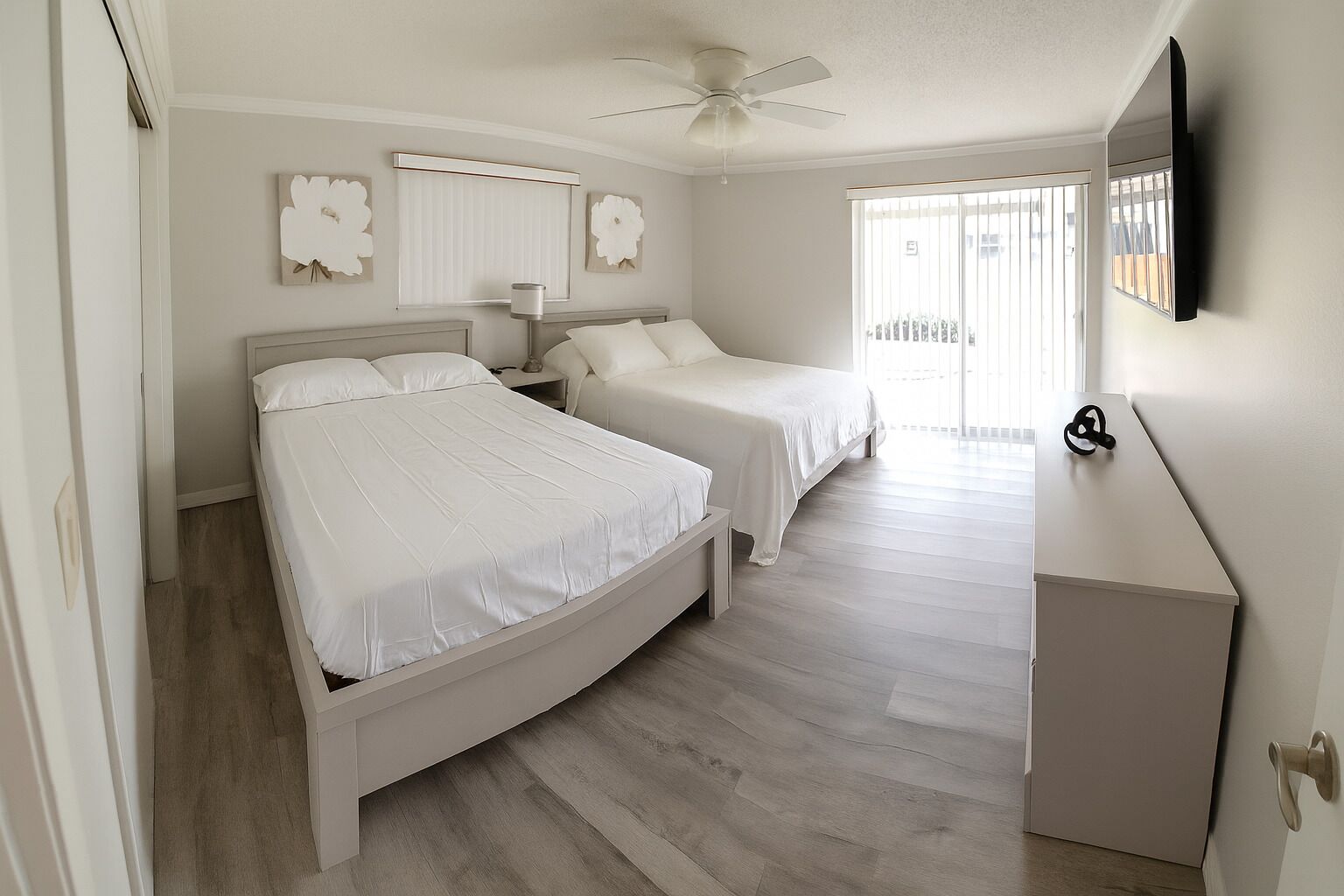 A bedroom with two beds, a ceiling fan, a bedside table with a lamp, and a TV mounted on the wall.