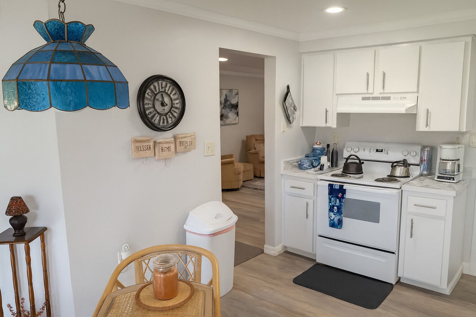 Una cocina moderna con armarios blancos, una estufa y una cafetera. Se observa un accesorio de luz decorativo azul y un reloj en la pared.
