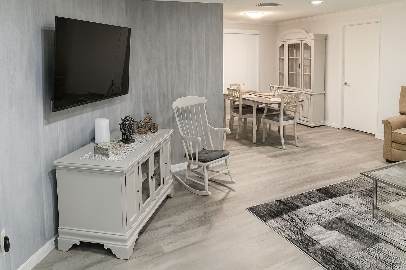 Een eethoek met een tafel en stoelen, een aan de muur bevestigde televisie, en een witte kast met decoratieve objecten.