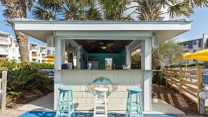 Un petit bar en plein air avec deux tabourets, une enseigne indiquant « Latte's 34 », et une vue sur des palmiers et des bâtiments en arrière-plan.